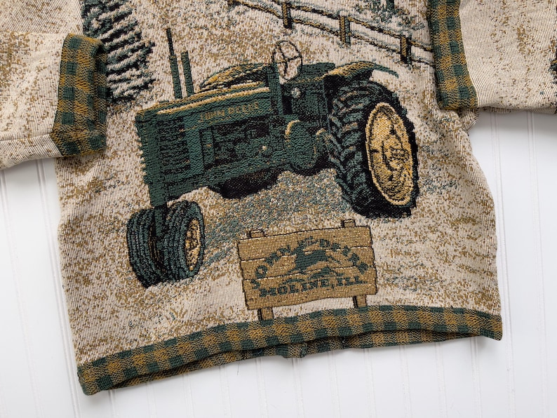 Puede incluir: Un su&eacute;ter beige con un dise&ntilde;o tejido verde y marr&oacute;n de un tractor John Deere. El su&eacute;ter tiene un ribete de cuadros verde y marr&oacute;n alrededor de los bordes. El tractor tiene el texto "John Deere" y "Moline, Ill." en &eacute;l.