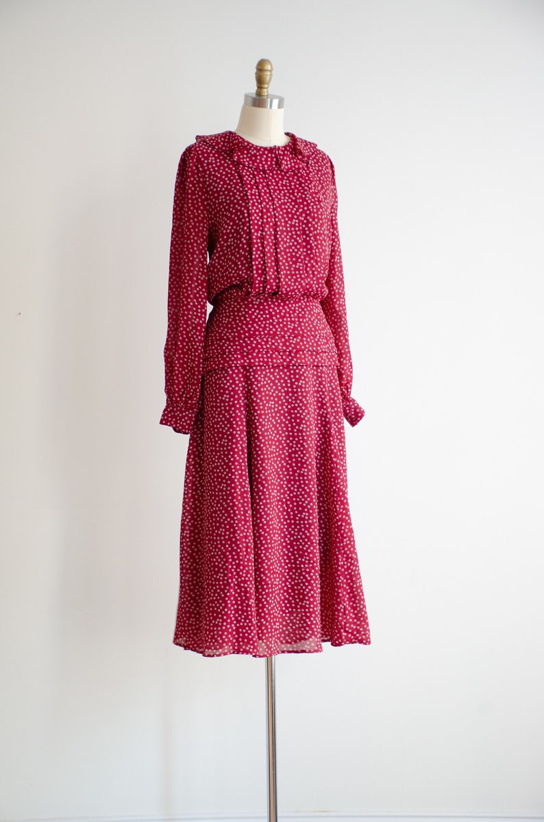 May include: A red dress with a white floral pattern. The dress has a ruffled collar and long sleeves. It is made of a lightweight fabric and has a flowing skirt.