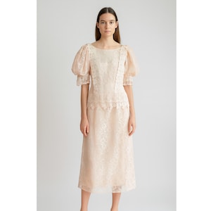 May include: A light peach-colored dress with a floral lace pattern. The dress features short, puffy sleeves and a midi-length skirt. The neckline is a wide, rounded scoop. The dress is made of a sheer fabric, giving it a delicate appearance.