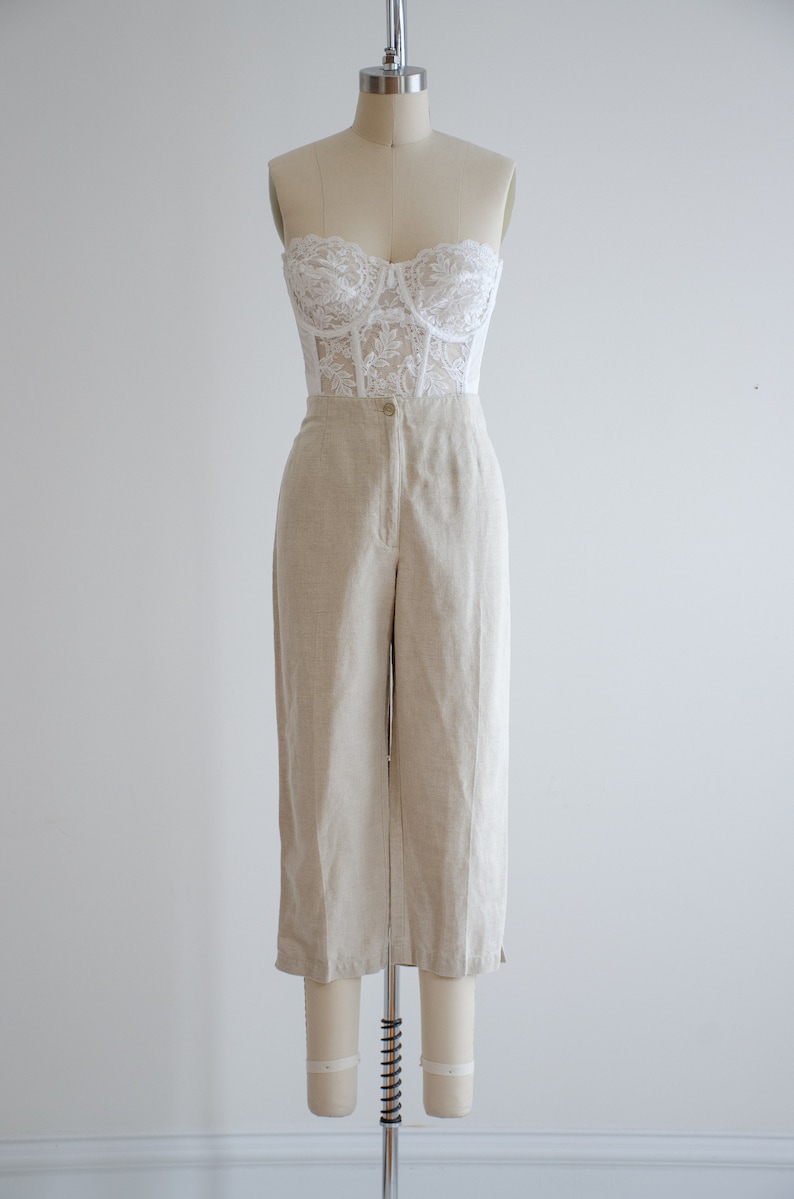 Puede incluir: Un top de cors&eacute; de encaje blanco combinado con pantalones de lino beige de cintura alta.