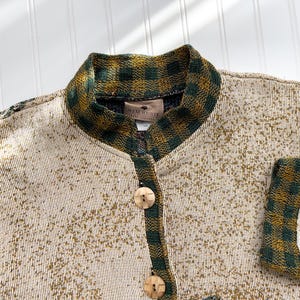 Puede incluir: Un c&aacute;rdigan de color crema con un ribete de cuadros verdes y marrones alrededor del cuello y las mangas. El c&aacute;rdigan tiene cierre de botones y la etiqueta "New Heather" es visible en el interior del cuello.