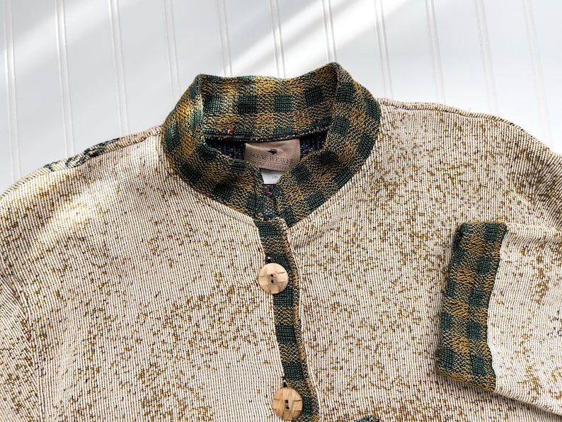 Puede incluir: Un c&aacute;rdigan de color crema con un ribete de cuadros verdes y marrones alrededor del cuello y las mangas. El c&aacute;rdigan tiene cierre de botones y la etiqueta "New Heather" es visible en el interior del cuello.