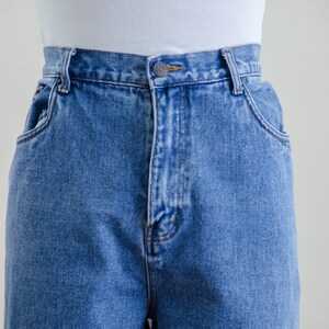 May include: Close-up of light blue denim jeans with a button closure and front pockets. The jeans have a classic, high-waisted design. A white ribbed tank top is visible above the jeans.