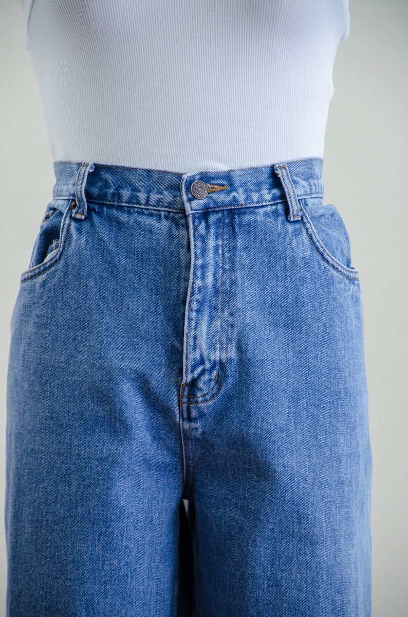 May include: Close-up of light blue denim jeans with a button closure and front pockets. The jeans have a classic, high-waisted design. A white ribbed tank top is visible above the jeans.