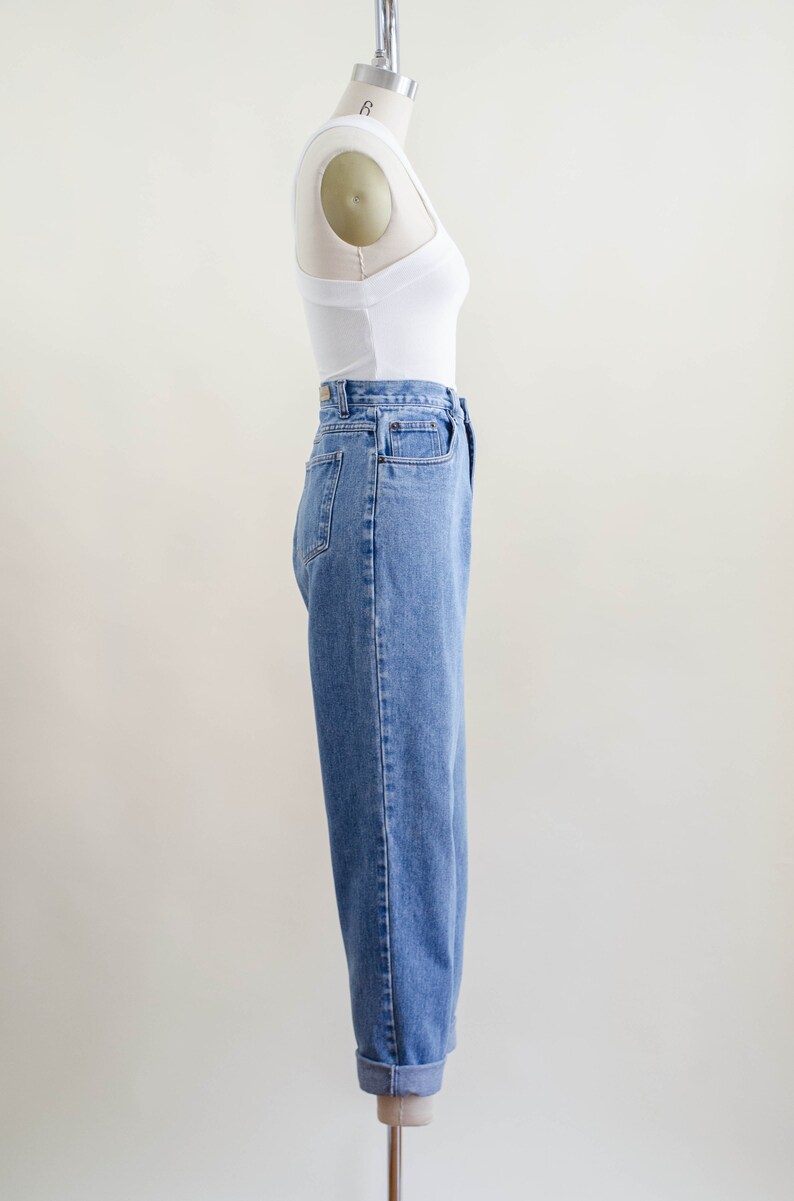 May include: A side view of a white tank top and light blue denim jeans on a mannequin. The jeans have a high waist and a relaxed fit, with the cuffs rolled up. The tank top is a simple, fitted style.