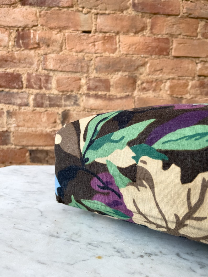 May include: A close-up of a brown, beige, green, and purple floral patterned fabric cushion. The cushion is sitting on a white marble surface.