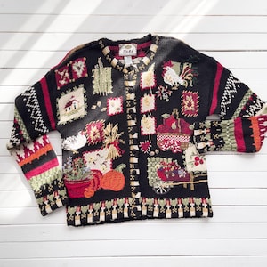May include: Black cardigan sweater with colorful embroidered patches featuring farm scenes, pumpkins, and a rooster. The sweater has a button-up front and long sleeves with patterned cuffs. The brand tag is visible at the neckline.
