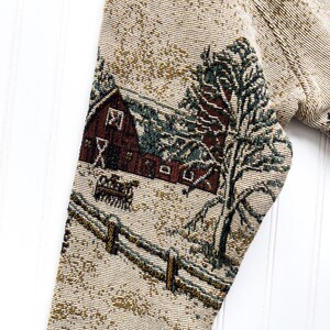 Puede incluir: Un su&eacute;ter beige con un dise&ntilde;o de tapiz tejido de una escena de invierno. La escena presenta un granero rojo, un &aacute;rbol verde y un campo cubierto de nieve con una cerca de madera.