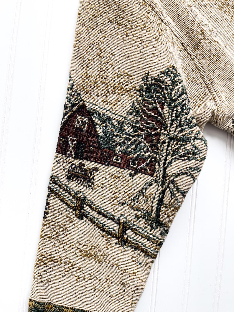 Puede incluir: Un su&eacute;ter beige con un dise&ntilde;o de tapiz tejido de una escena de invierno. La escena presenta un granero rojo, un &aacute;rbol verde y un campo cubierto de nieve con una cerca de madera.