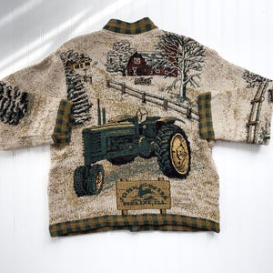 Puede incluir: Un c&aacute;rdigan beige con un tractor verde y marr&oacute;n y una escena de granja tejida en la tela. El c&aacute;rdigan tiene un cuello y pu&ntilde;os a cuadros. El texto "John Deere" y "Moline, Ill." est&aacute; en un letrero en la imagen.