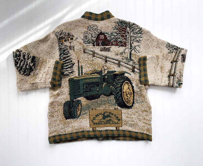 Puede incluir: Un c&aacute;rdigan beige con un tractor verde y marr&oacute;n y una escena de granja tejida en la tela. El c&aacute;rdigan tiene un cuello y pu&ntilde;os a cuadros. El texto "John Deere" y "Moline, Ill." est&aacute; en un letrero en la imagen.