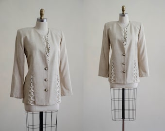 Puede incluir: Una chaqueta beige con un detalle de encaje blanco. La chaqueta tiene un cuello de muesca, mangas largas y un cierre de un solo botón.