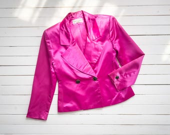 May include: A vibrant fuchsia satin blazer with a double-breasted design. The jacket features a notched collar, long sleeves, and black buttons. The garment is laid flat on a white wooden surface.