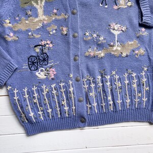 May include: A light blue knitted cardigan with embroidered floral designs, including a bicycle, a tree, and a bird. The cardigan has a button-up front and a ribbed hem.
