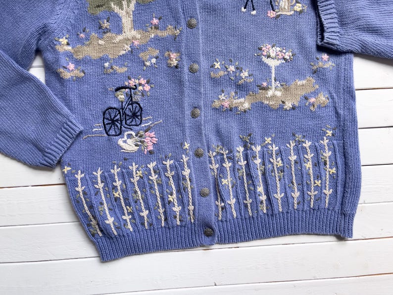 May include: A light blue knitted cardigan with embroidered floral designs, including a bicycle, a tree, and a bird. The cardigan has a button-up front and a ribbed hem.