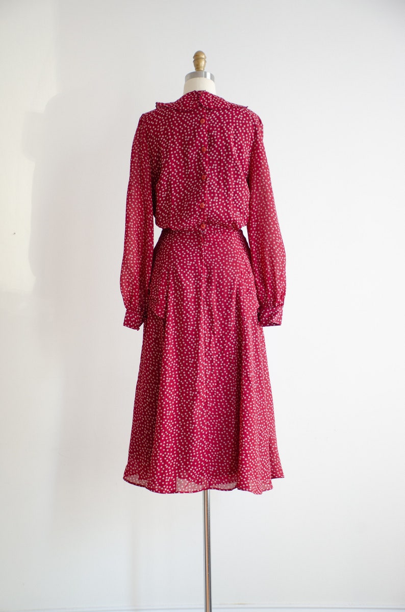 May include: A long-sleeved, red dress with a white floral pattern. The dress has a ruffled collar and a button-up closure in the back.