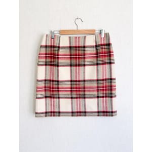 May include: A knee-length skirt featuring a plaid pattern in cream, red, black, and grey. The skirt is suspended from a wooden hanger with two metal clips attached to the waistband, showcasing a straight-cut design with a visible woven texture.