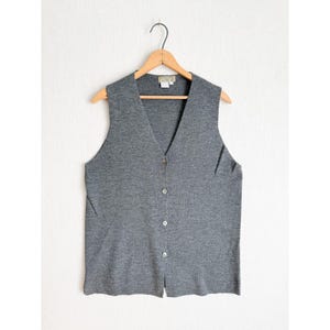 Puede incluir: Un chaleco gris con botones y cuello en V. El chaleco sin mangas cuelga de una percha de madera. Los botones son de color claro y la tela parece ser de punto. El chaleco es de color liso y tiene un estilo sencillo y clásico.
