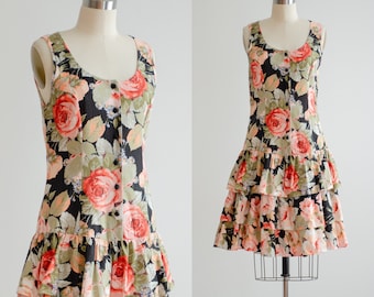 Könnte beinhalten: Ein schwarzes, ärmelloses Kleid mit einem Blumendruck mit rosa und orangefarbenen Rosen. Das Kleid hat einen Knopfleisten vorne und einen Stufenrock.