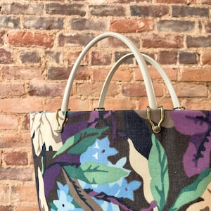 May include: A white canvas tote bag with double handles and a floral print featuring blue, green, and purple flowers on a brown background.