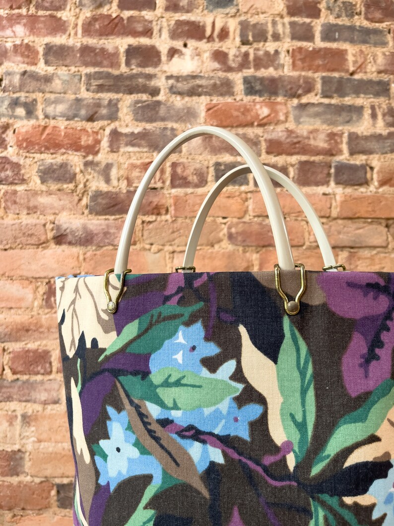 May include: A white canvas tote bag with double handles and a floral print featuring blue, green, and purple flowers on a brown background.