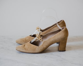 May include: A pair of light brown suede heeled shoes with a gold chain detail on the strap.