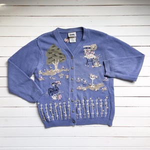May include: A light blue cardigan with embroidered floral and nature scenes. The cardigan has a button-up front and long sleeves.