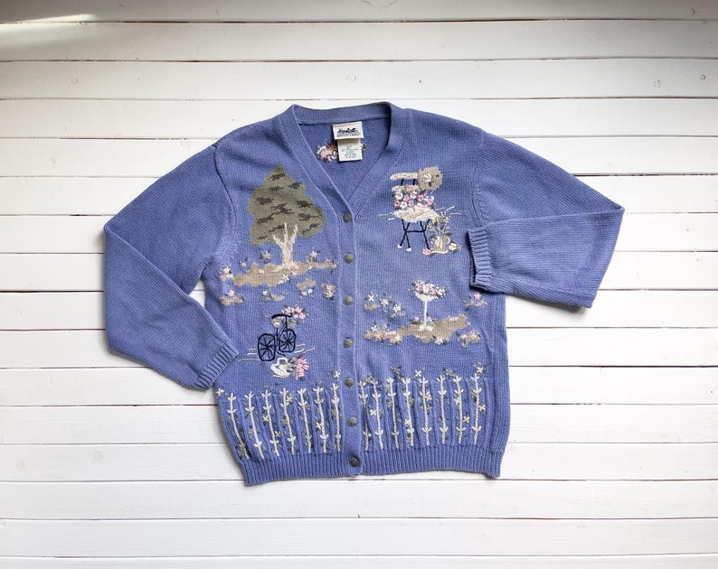 May include: A light blue cardigan with embroidered floral and nature scenes. The cardigan has a button-up front and long sleeves.