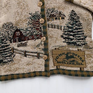 Puede incluir: Un c&aacute;rdigan beige con una escena de invierno tejida en la tela. La escena incluye una granja, &aacute;rboles y un letrero que dice "John Deere Moline, Ill."