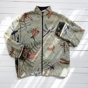 May include: A light green jacket with a high collar and a full-length black zipper. The jacket features a repeating pattern of dragonflies in shades of brown, blue, and pink. The sleeves have a decorative border.
