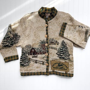 Puede incluir: Una chaqueta de punto color crema con ribetes de cuadros verdes y marrones. La chaqueta presenta una escena de invierno con un granero rojo, &aacute;rboles cubiertos de nieve y un letrero que dice "Town Deer, Molino, Ill."