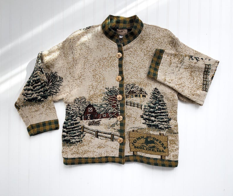 Puede incluir: Una chaqueta de punto color crema con ribetes de cuadros verdes y marrones. La chaqueta presenta una escena de invierno con un granero rojo, &aacute;rboles cubiertos de nieve y un letrero que dice "Town Deer, Molino, Ill."