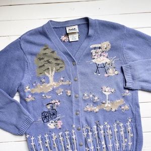 May include: A light blue knitted cardigan with embroidered floral designs, including a tree, a bicycle, and a table with flowers. The cardigan has a V-neck and button closure.