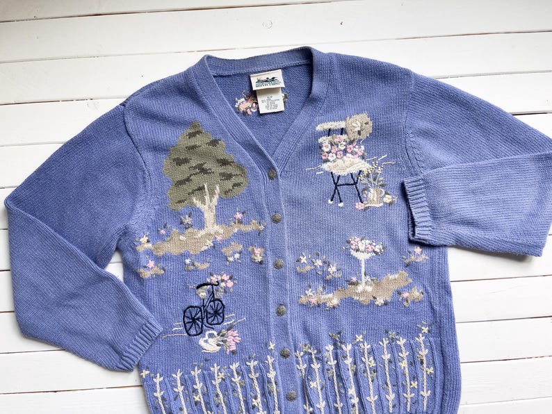 May include: A light blue knitted cardigan with embroidered floral designs, including a tree, a bicycle, and a table with flowers. The cardigan has a V-neck and button closure.