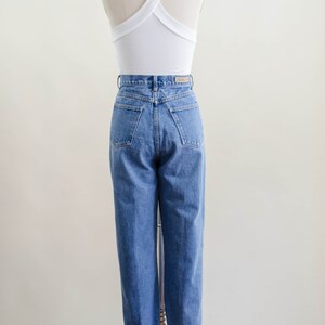 May include: A white halter top with a criss-cross back design paired with high-waisted, light-wash blue jeans. The jeans have a classic fit with a straight leg and cuffed hem. The outfit is displayed on a mannequin.