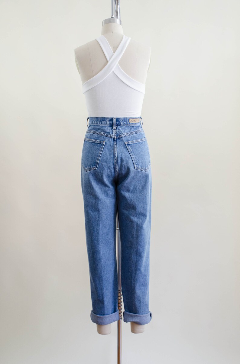 May include: A white halter top with a criss-cross back design paired with high-waisted, light-wash blue jeans. The jeans have a classic fit with a straight leg and cuffed hem. The outfit is displayed on a mannequin.
