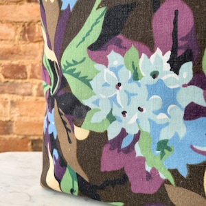 May include: A close-up of a brown fabric with a floral pattern. The fabric features large blue flowers with white centers and green leaves. The background is a dark brown with hints of purple.