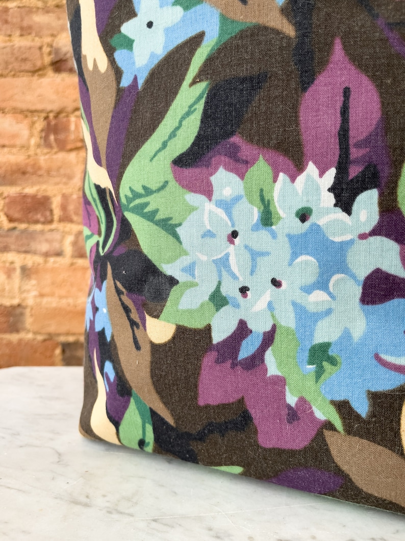 May include: A close-up of a brown fabric with a floral pattern. The fabric features large blue flowers with white centers and green leaves. The background is a dark brown with hints of purple.