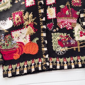 May include: Black cardigan sweater with colorful embroidered autumn-themed patches. The patches include pumpkins, wheat, flowers, and a basket of fruit. The sweater has a button-up front and a patterned border at the hem and cuffs.