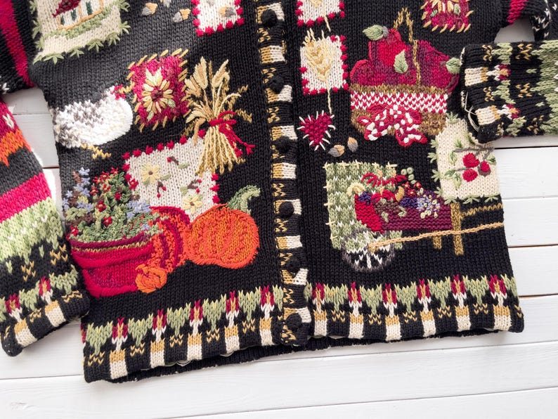 May include: Black cardigan sweater with colorful embroidered autumn-themed patches. The patches include pumpkins, wheat, flowers, and a basket of fruit. The sweater has a button-up front and a patterned border at the hem and cuffs.