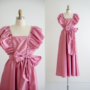 May include: A pink satin dress with a square neckline and ruffled sleeves. The dress has a large bow at the waist and a full skirt.