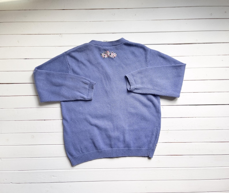 May include: A light blue knitted cardigan with a floral embroidery design on the back.