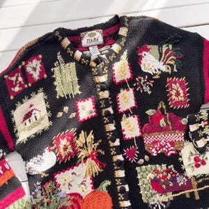 May include: Black button-up cardigan sweater with a variety of embroidered patches. The patches feature images of a rooster, pumpkins, wheat, and a house. The sleeves have a red and white geometric pattern. The label reads "TIARA".