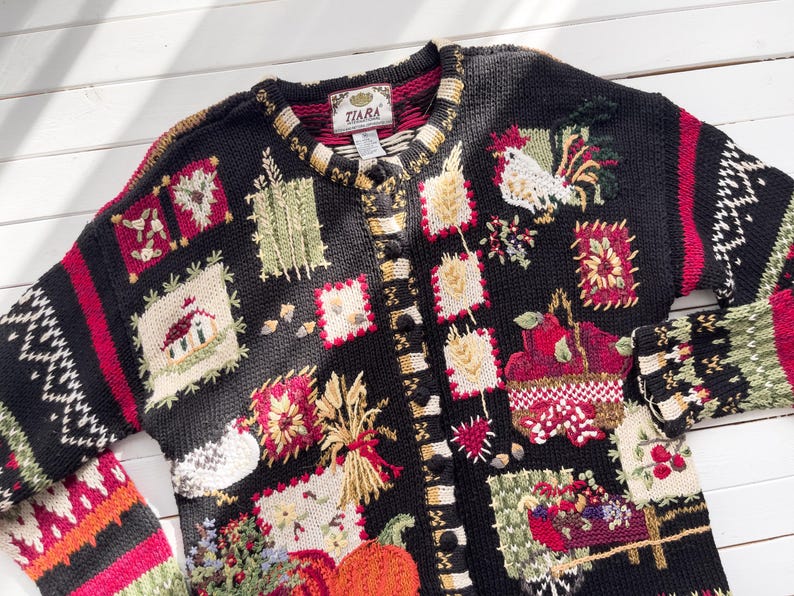 May include: Black button-up cardigan sweater with a variety of embroidered patches. The patches feature images of a rooster, pumpkins, wheat, and a house. The sleeves have a red and white geometric pattern. The label reads "TIARA".