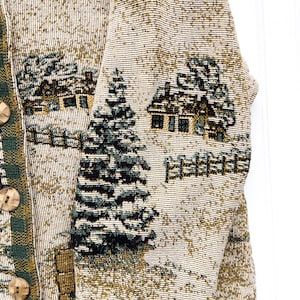 Puede incluir: Una chaqueta de punto de color crema con ribetes de cuadros verdes y marrones. La chaqueta presenta un dise&ntilde;o tejido de casas, &aacute;rboles y vallas.