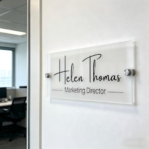 May include: A clear acrylic office sign with the name "Helen Thomas" in a stylish script font and the title "Marketing Director" in a simple sans-serif font. The sign is mounted on a white wall with silver-colored hardware.