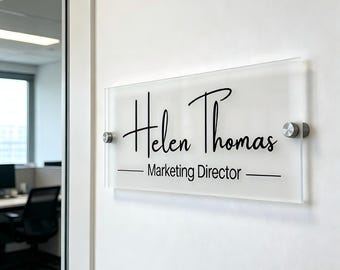 Puede incluir: Un letrero de oficina de acrílico transparente con el nombre "Helen Thomas" en una fuente elegante y el título "Marketing Director" en una fuente sencilla sin serifa. El letrero está montado en una pared blanca con herrajes plateados.