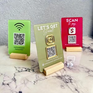May include: Three acrylic business signs in green, olive green, and red, each mounted on a wooden base. The green sign displays a WiFi symbol and QR code. The olive green sign says "LET'S GET Social!" with an Instagram logo and QR code. The red sign says "SCAN to pay" with a dollar sign and QR code.