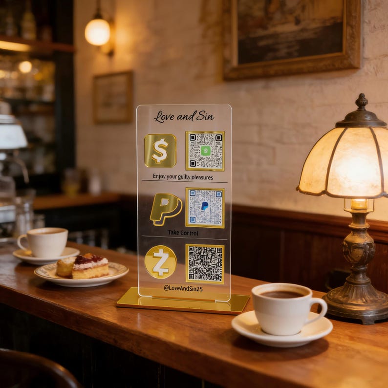 Op de afbeelding: Een helder acryl bord met gouden accenten toont betalingsopties en QR-codes. Het bord bevat de tekst "Love and Sin", "Enjoy your guilty pleasures" en "Take Control". Een koffiekopje en een dessertbord staan op een houten oppervlak.