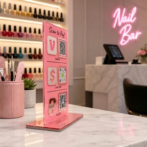 Puede incluir: Un letrero de acr&iacute;lico rosa con el texto "Scan to Pay!" y opciones de pago: Venmo, Cash App y PayPal, cada uno con un c&oacute;digo QR. El letrero est&aacute; sobre una superficie de m&aacute;rmol, con botellas de esmalte de u&ntilde;as en el fondo. Un letrero de ne&oacute;n rosa dice "Nail Bar".